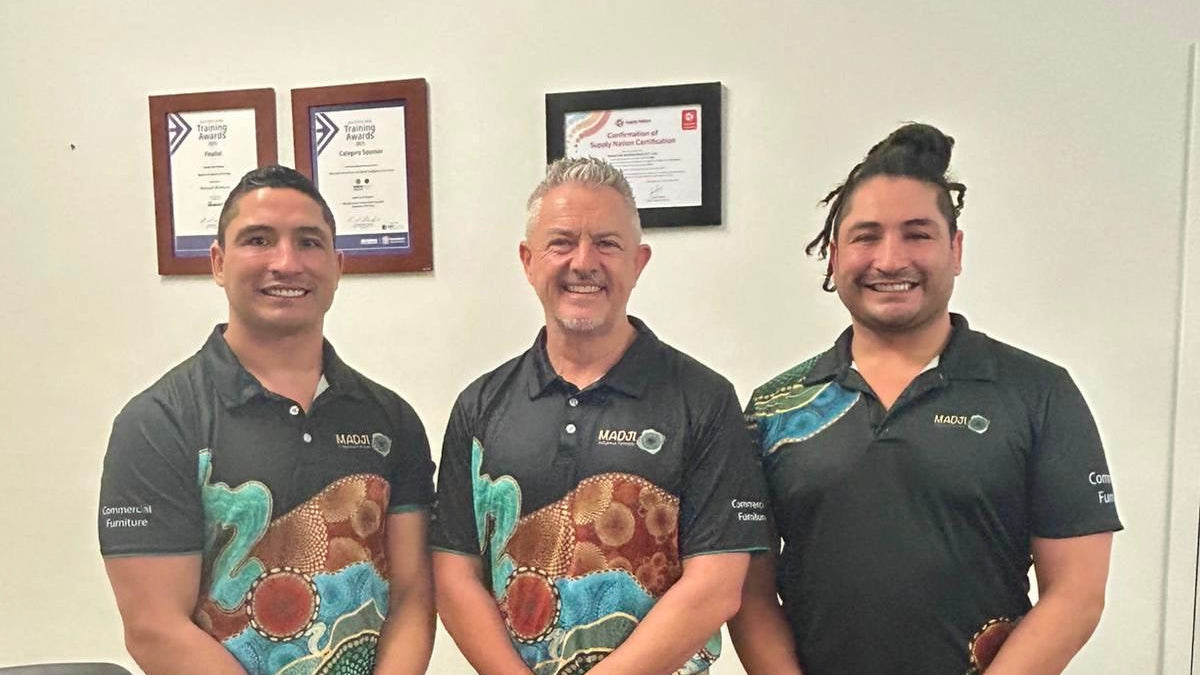 Three men in matching shirts standing in an office setting with framed certificates on the wall.