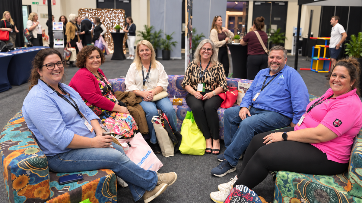 A group of seven people sitting together on an yarning circle at a conference or event, with booths and other attendees visible in the background.