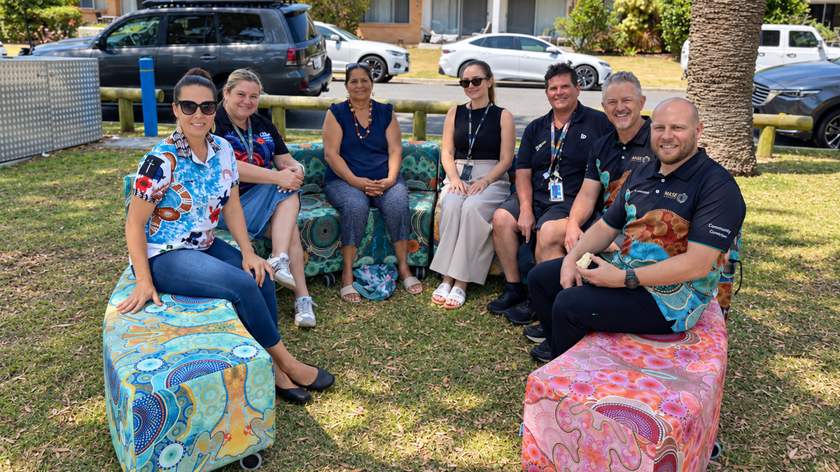 Seven people sitting outdoors on colorful indigenous ottomans in a park-like setting with apartment buildings, parked cars, and a palm tree in the background.