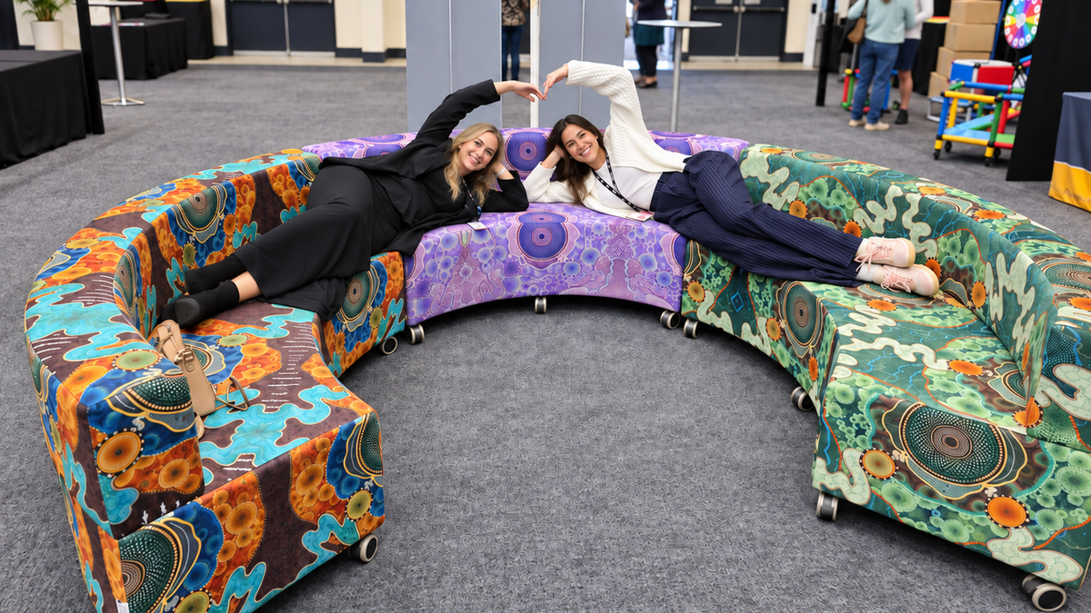 Two School Business managers reclining on a yarning circle chair with an Indigenous artwork, orange, purple, and green, set in a conference center hall.